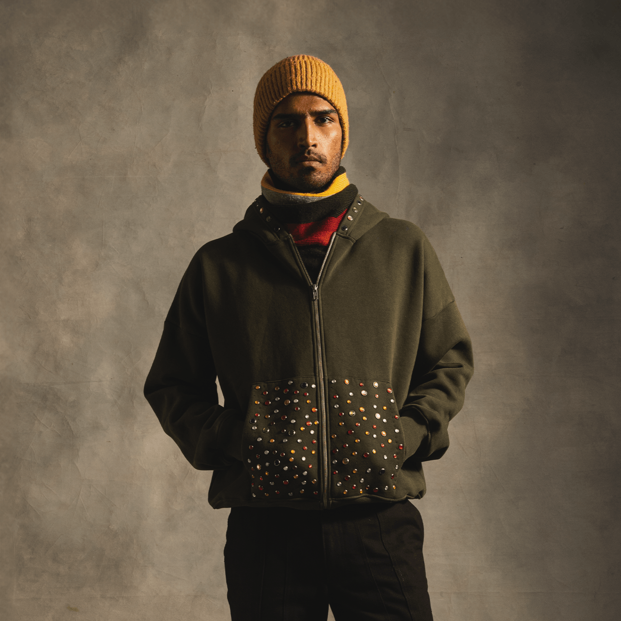Man wearing olive green studded streetwear hoodie, yellow knit beanie, and black pants
