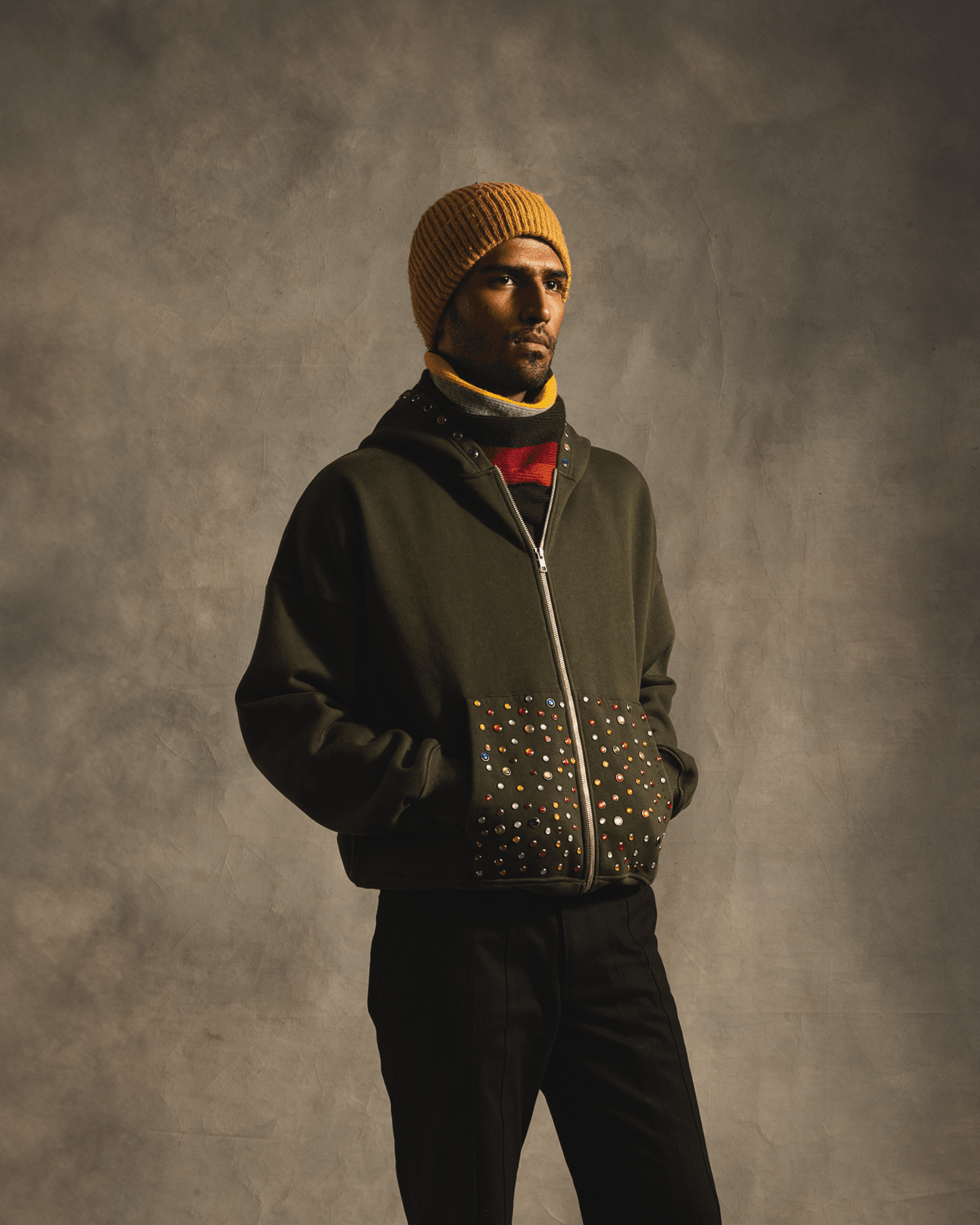 Man wearing dark green hoodie with multicolored studded pockets and mustard beanie