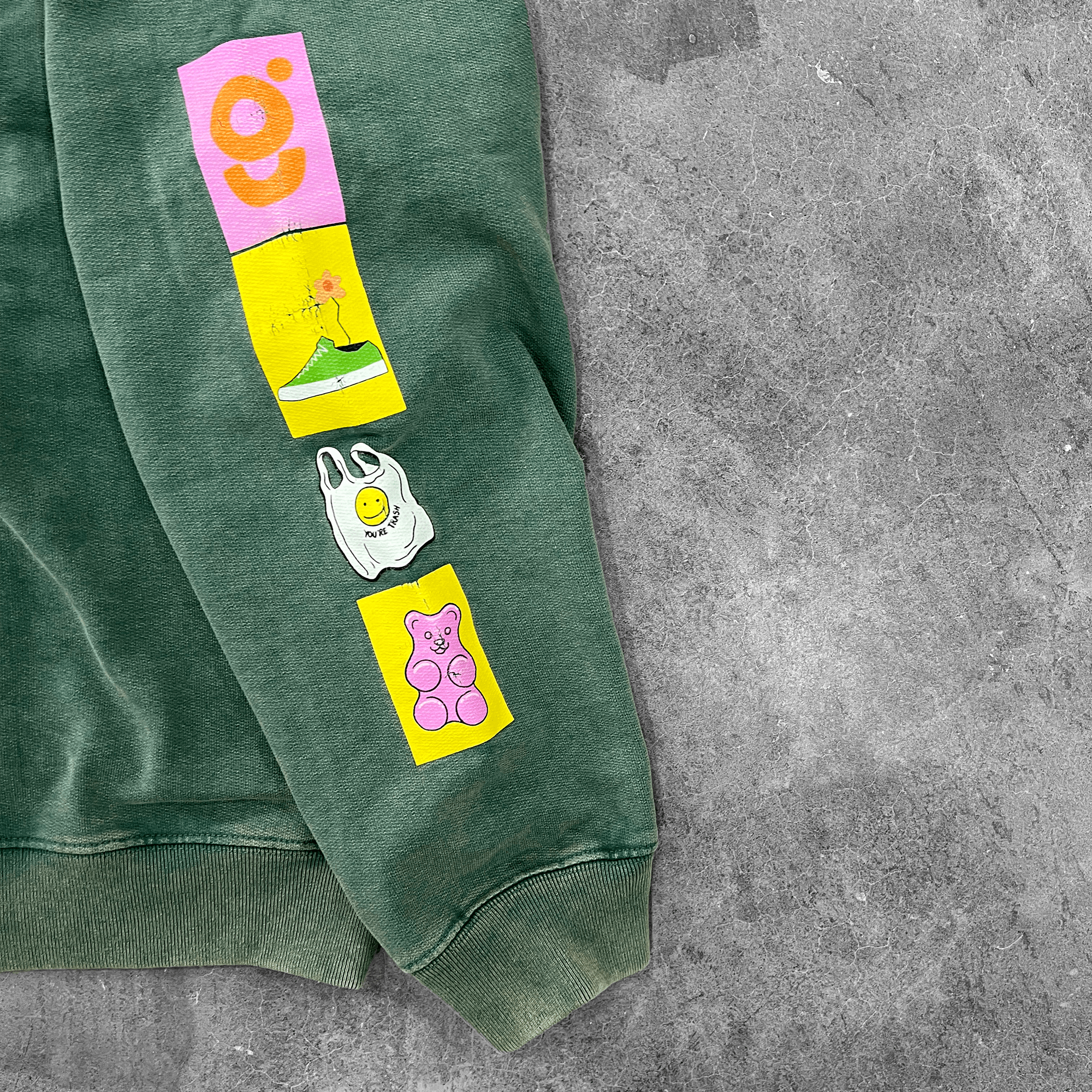 Green streetwear hoodie sleeve with colorful graphic patches on concrete background