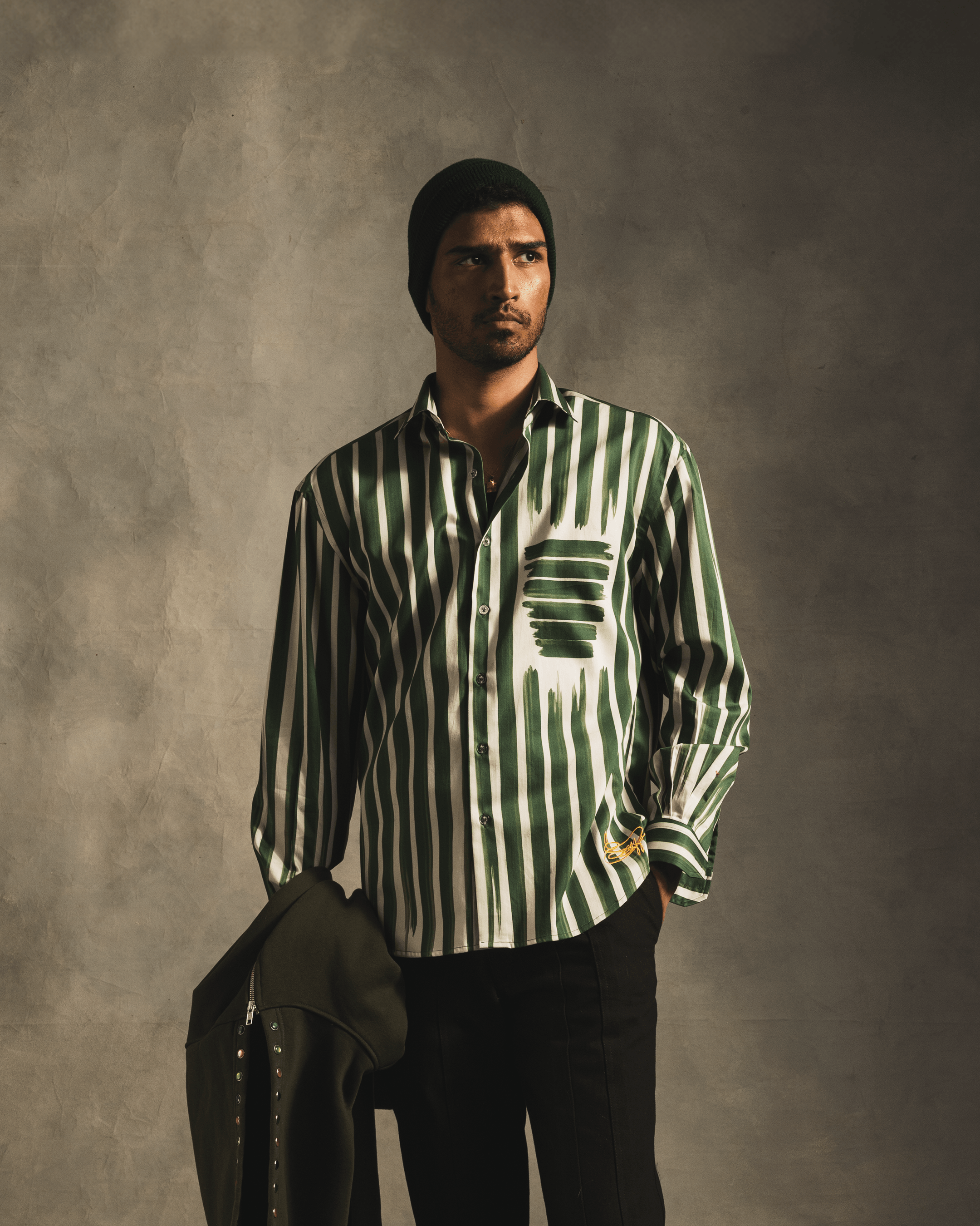 Man wearing green and white striped button-up shirt and black beanie, holding jacket