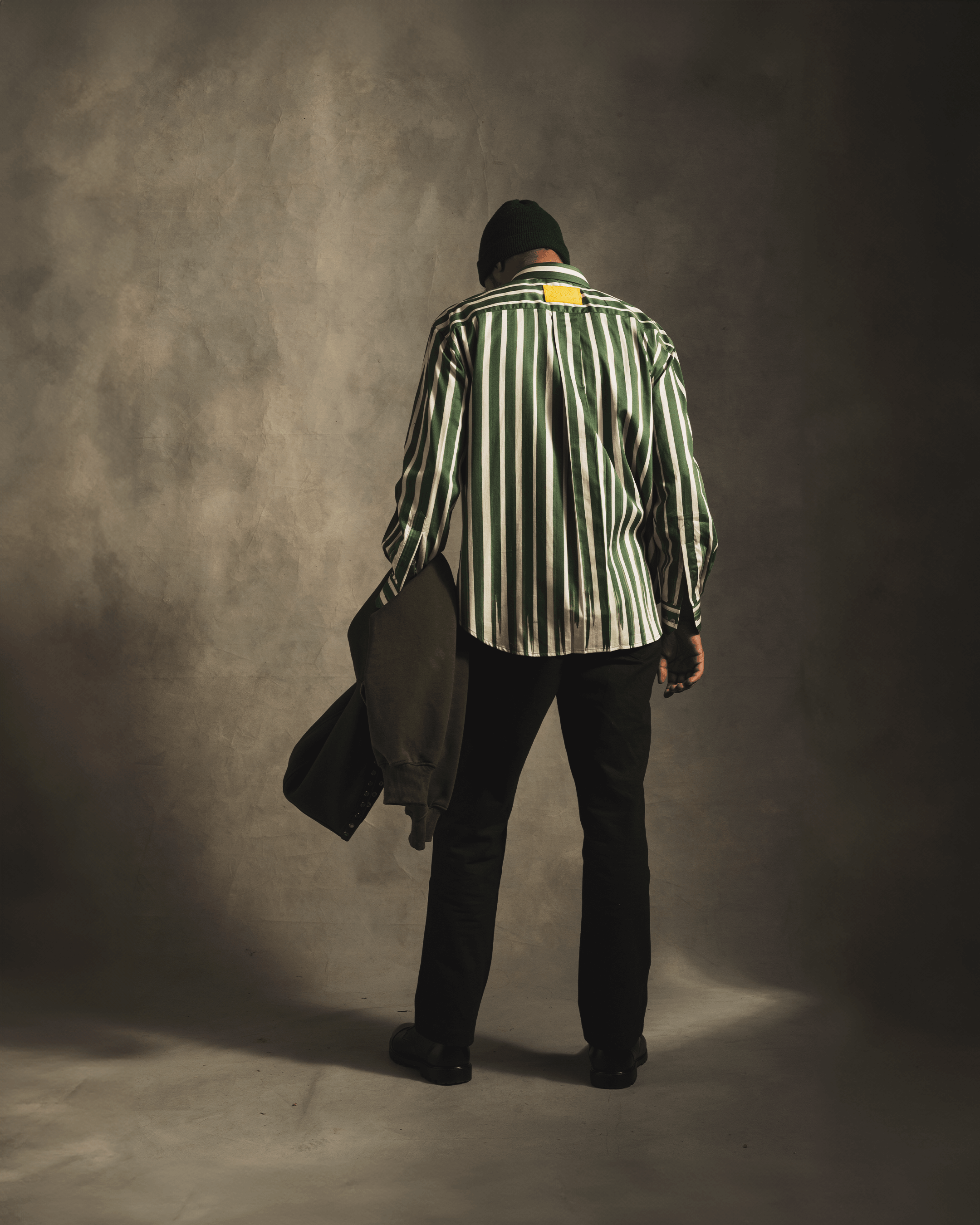 Man in green striped shirt, black pants, beanie, holding jacket, streetwear fashion