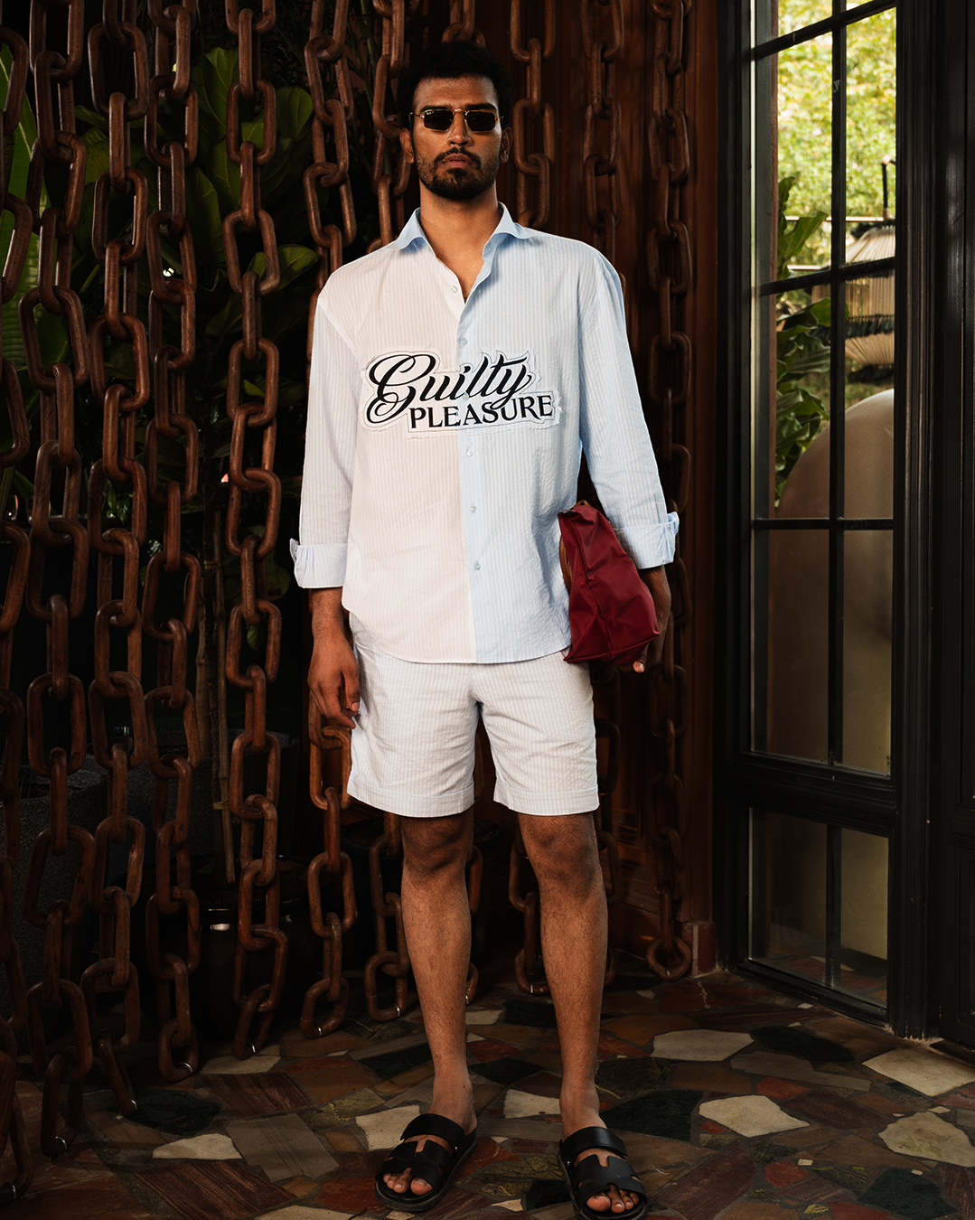 Man wearing striped 'Guilty Pleasure' shirt and shorts, streetwear style, holding red bag.