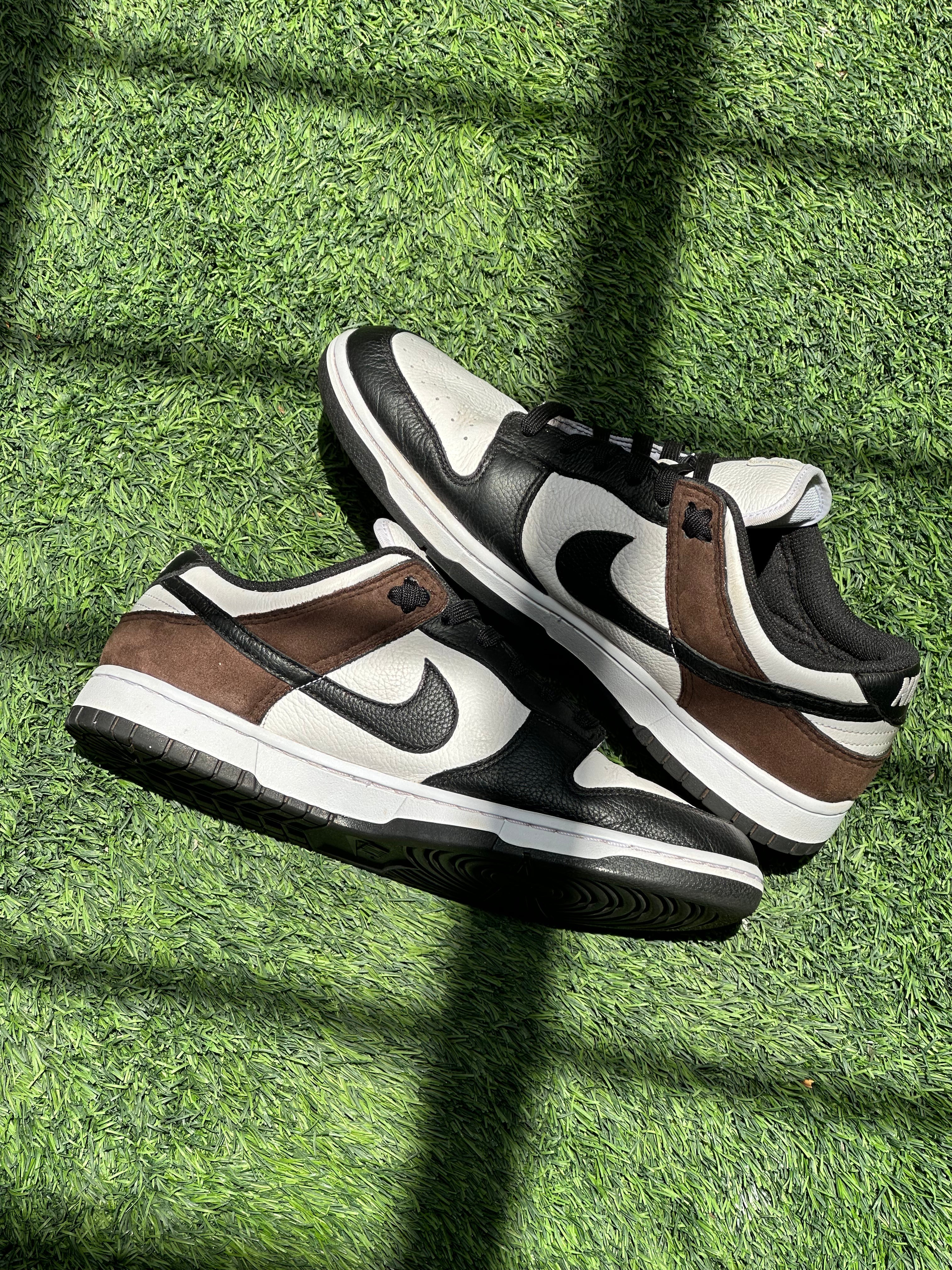 Black, white, and brown Nike Dunk Low sneakers on green artificial grass with sunlight shadows.
