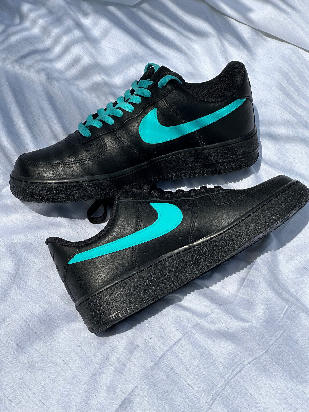 Black low-top sneakers with teal swoosh and teal laces on white fabric background.