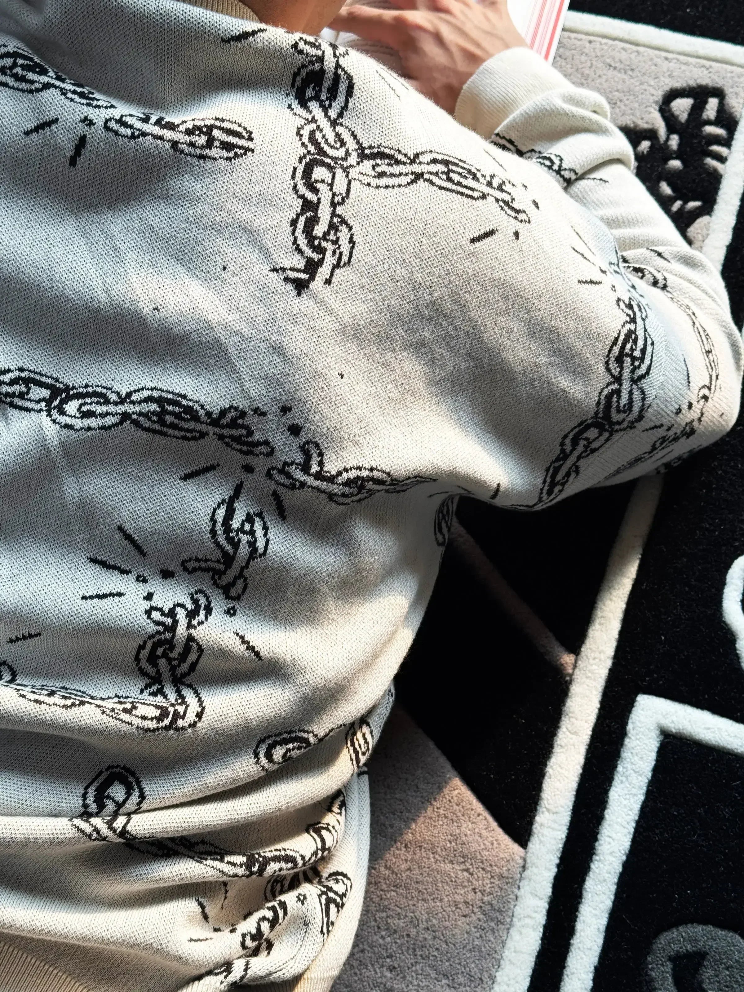 Person wearing light gray sweater with black broken chain graphic design lying on patterned carpet