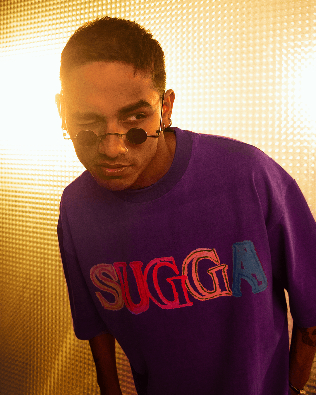 Man in purple streetwear t-shirt with colorful SUGGA text, wearing round sunglasses, bold lighting