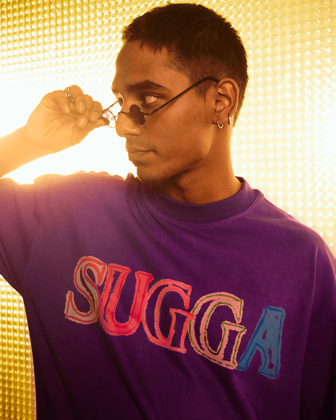 Man wearing purple streetwear t-shirt with colorful SUGGA text, trendy sunglasses, gold backdrop
