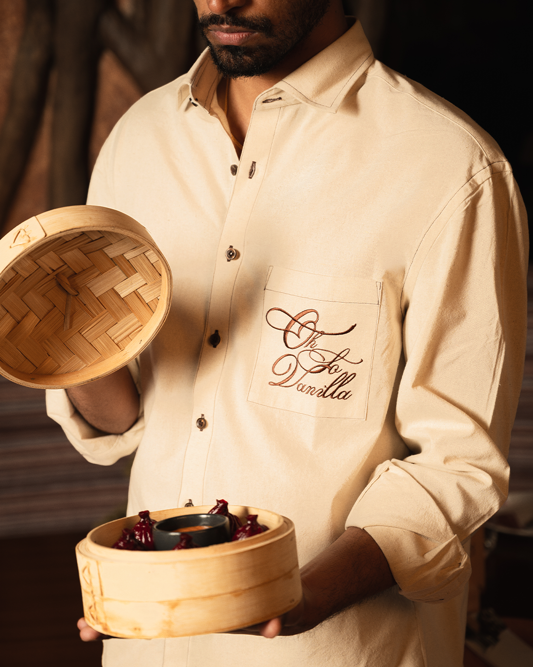Man in beige shirt with 'Oh So Vanilla' embroidery holding bamboo steamer basket with food