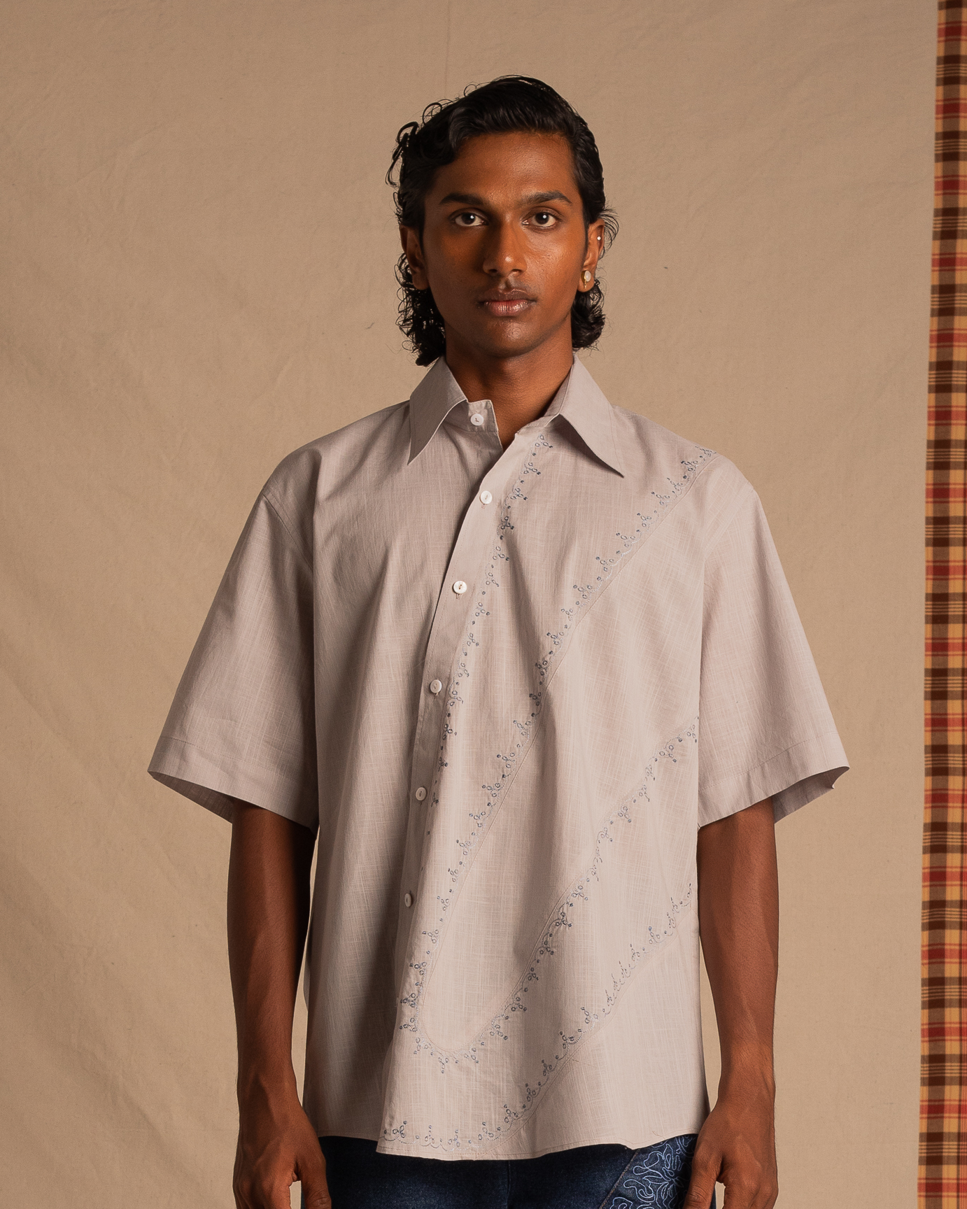 Man wearing oversized short sleeve light gray button-up shirt with subtle embroidered pattern, standing against neutral backdrop.