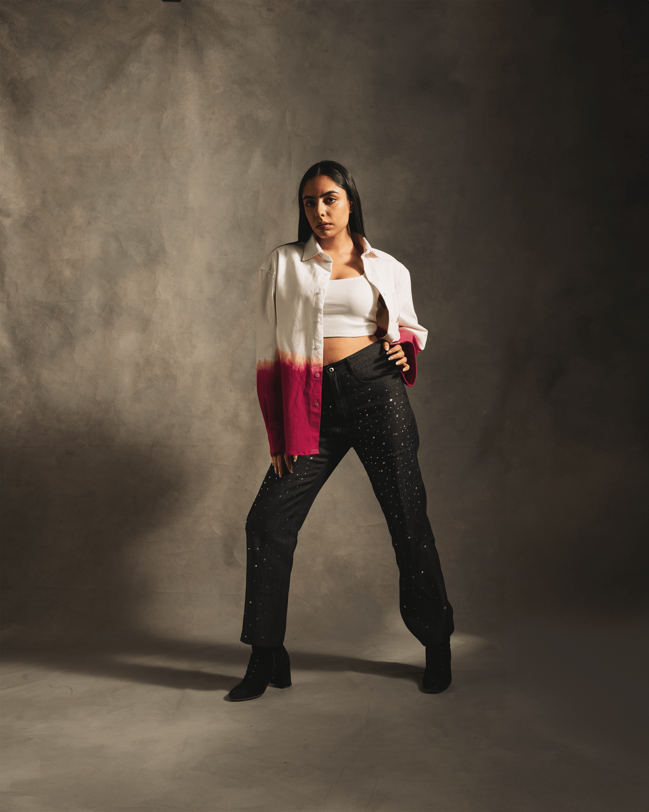 Woman in crop top, white and pink ombré jacket, and black studded jeans posing indoors
