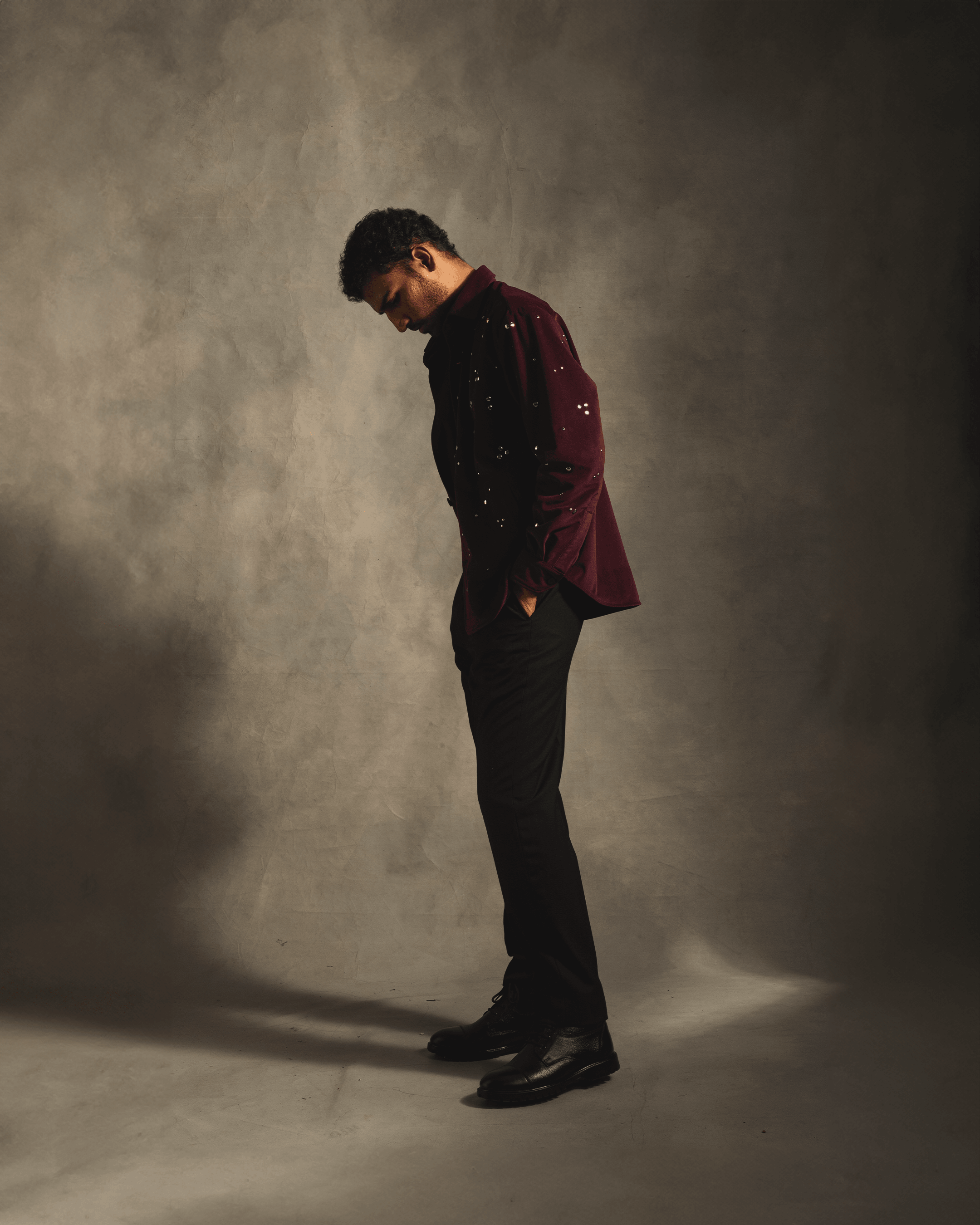 Man wearing a red velvet studded shirt with black pants and boots in a studio setting