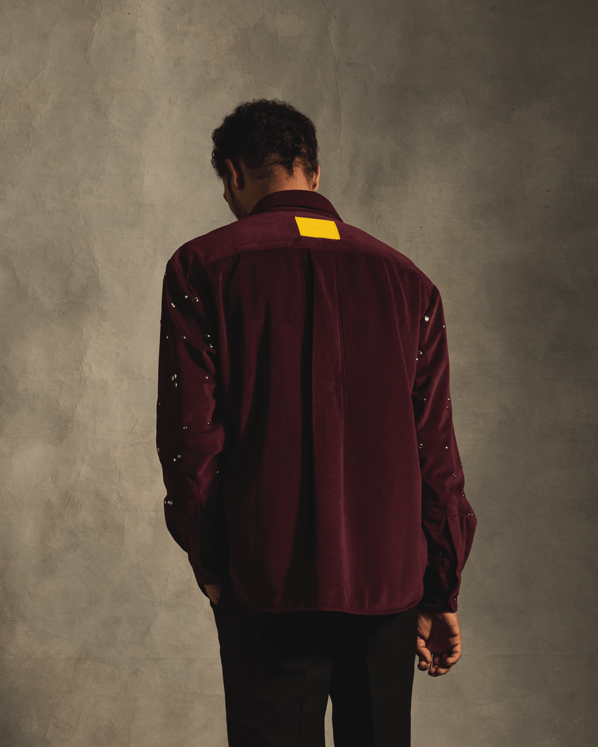 Man wearing a red velvet long sleeve shirt with studded sleeves, standing against a gray wall