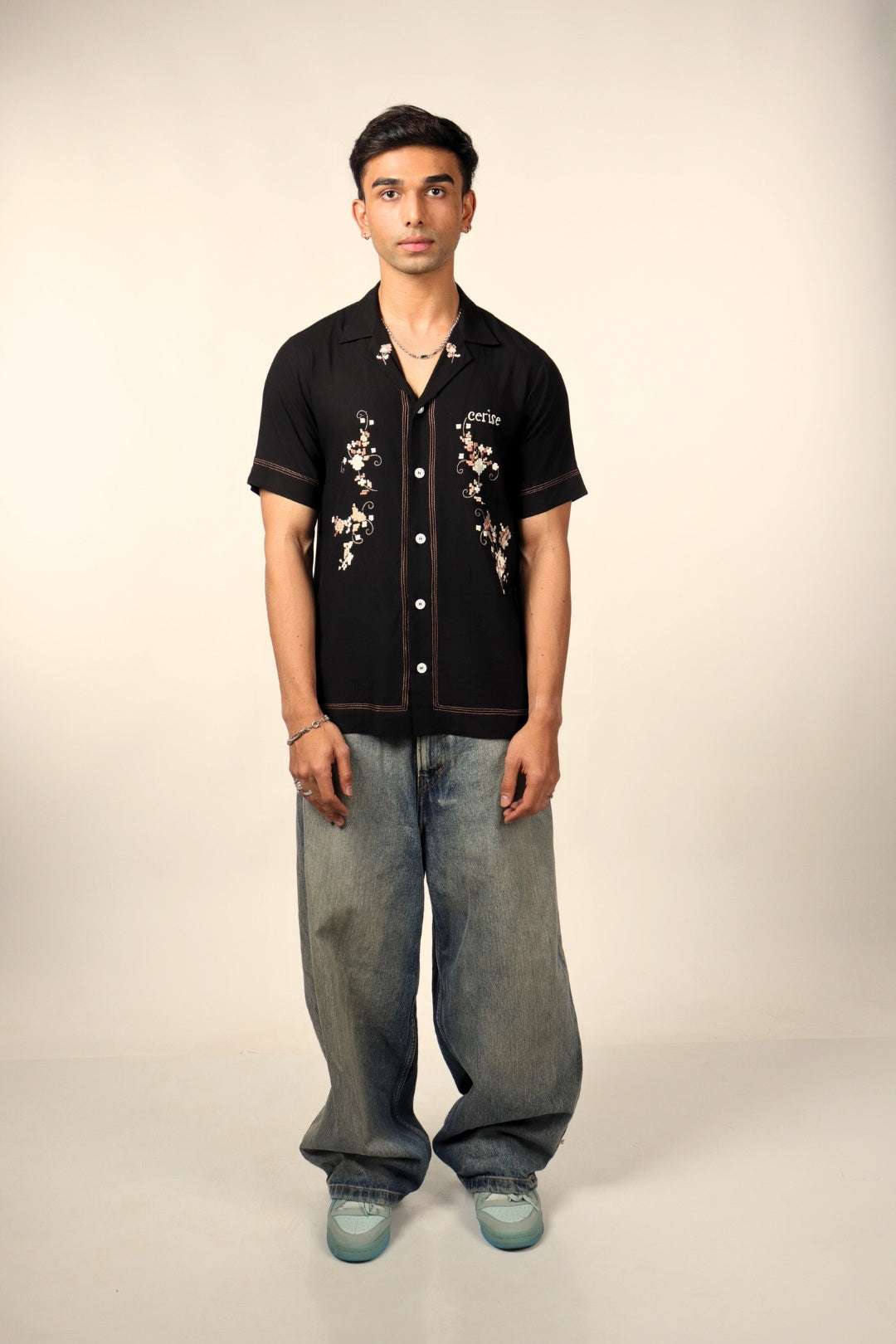 Man in floral embroidered black short sleeve shirt, baggy jeans, and light blue sneakers standing against beige background