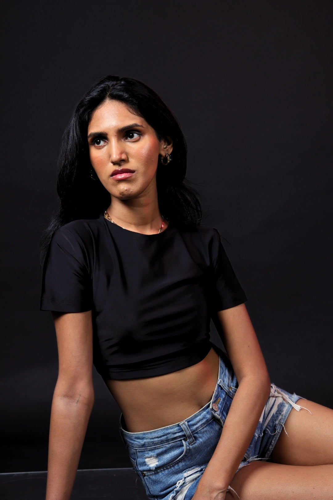Woman in black crop top and distressed denim shorts posing against dark background, streetwear fashion