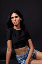 Woman in black crop top and distressed denim shorts posing against dark background, streetwear fashion