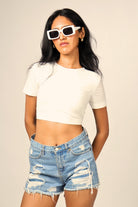 Woman wearing white crop top, distressed denim shorts, and white sunglasses, streetwear fashion.