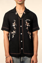 Man wearing a black short sleeve button-up shirt with floral embroidery and 'Cerise' text, paired with jeans.