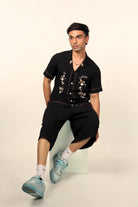 Man in black floral embroidered shirt, black shorts, and blue sneakers, streetwear style