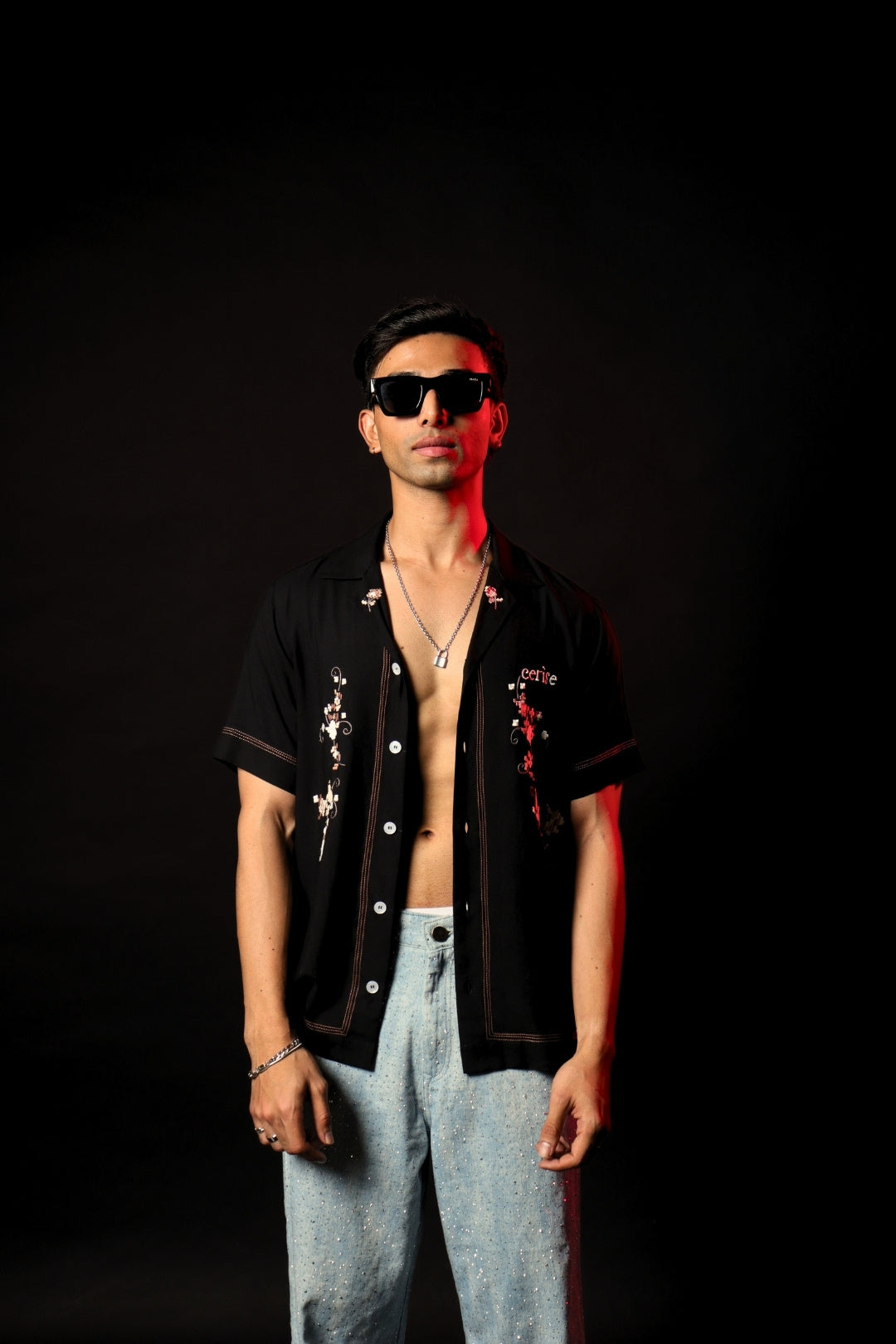 Man wearing black embroidered open shirt, blue jeans, and sunglasses in streetwear style