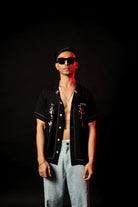 Man wearing black embroidered open shirt, blue jeans, and sunglasses in streetwear style