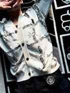 Person wearing a white cardigan with black chain pattern and black pants with a Lost Exit patch lying on a black and white patterned carpet