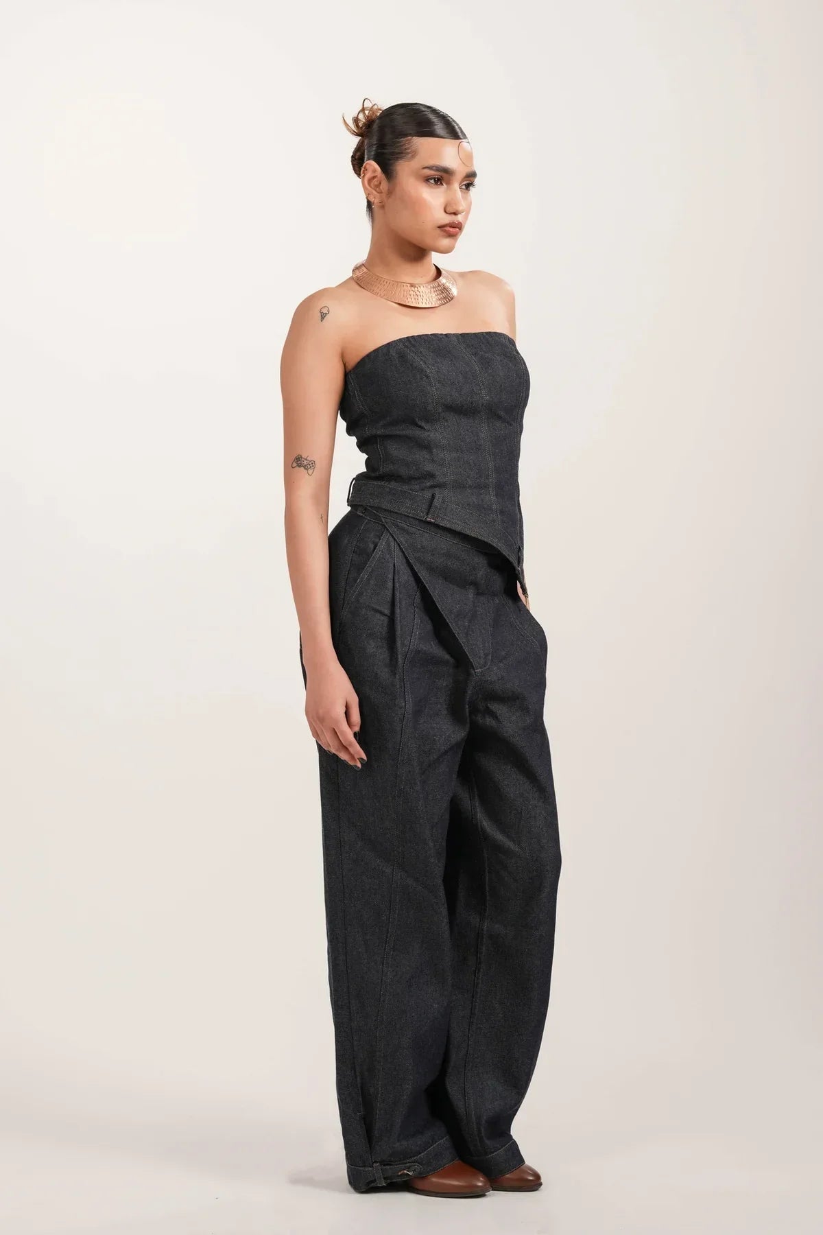 Woman wearing strapless structured black denim jumpsuit with wide legs and brown shoes on neutral background